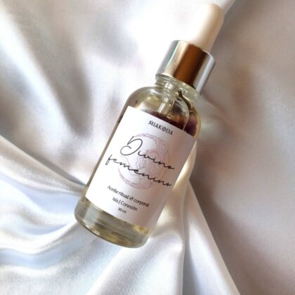 aceite cristalino con etiqueta blanca, dentro de el hay bulbos de rosas. Esta sobre una cama de satín blanco