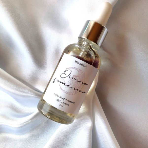 aceite cristalino con etiqueta blanca, dentro de el hay bulbos de rosas. Esta sobre una cama de satín blanco