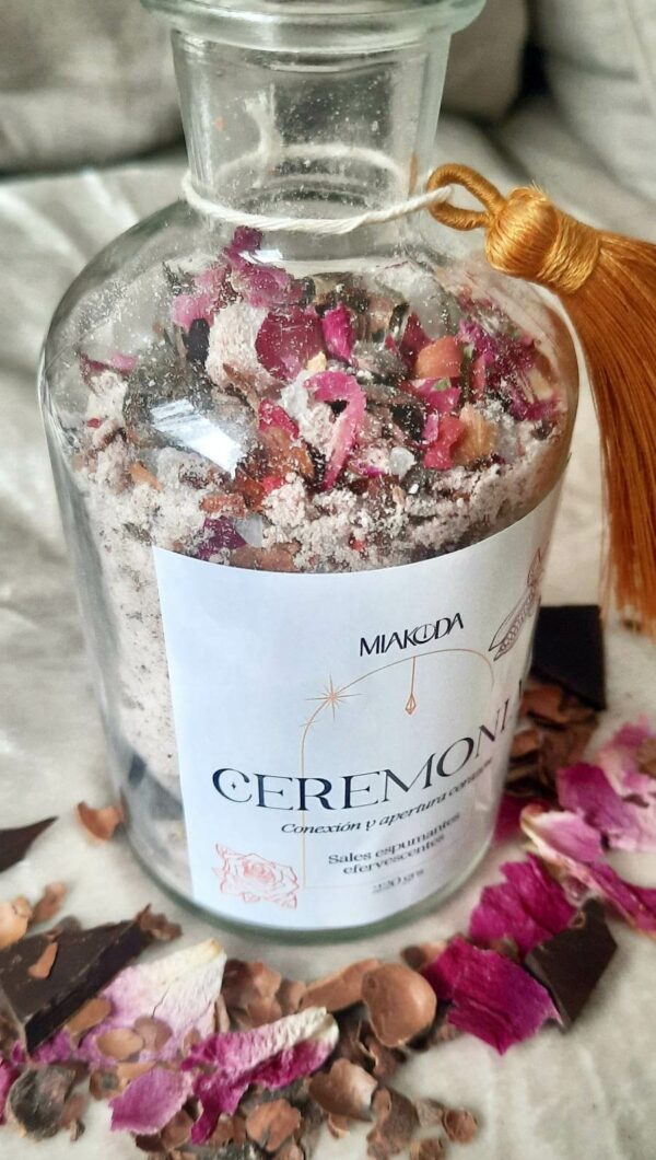 Frasco de vidrio relleno son sal de mar, rosas y cacao. Tiene una etiqueta blanca y rodeada de rosas y diferentes tipos de cacao